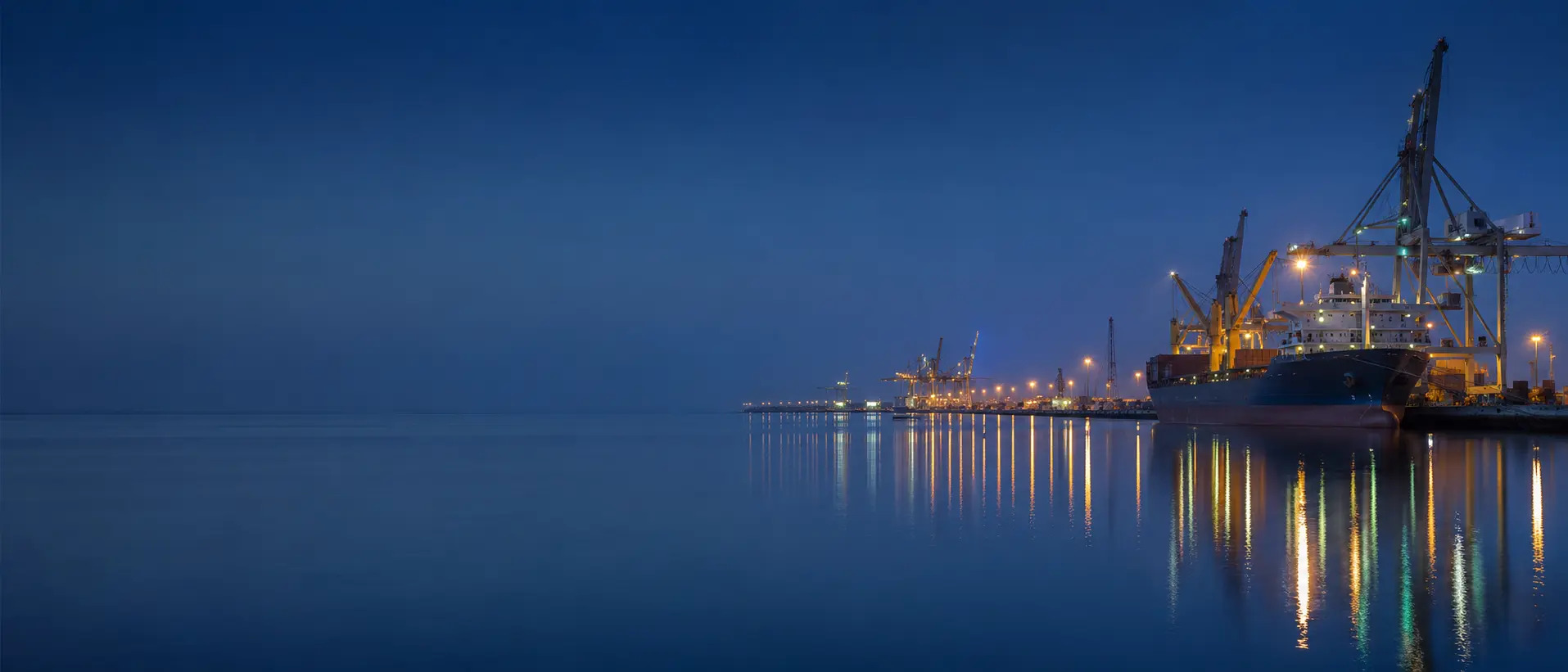 Commercial cargo vessel berthed under port cranes at blue hour.